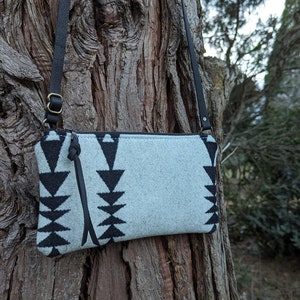 Puede incluir: Un bolso bandolera azul claro con un patr&oacute;n geom&eacute;trico negro. El bolso tiene una correa de cuero negro y un tirador de cremallera. El bolso est&aacute; colgado de un tronco de &aacute;rbol. El bolso mide aproximadamente 25 cm de ancho y 15 cm de alto.