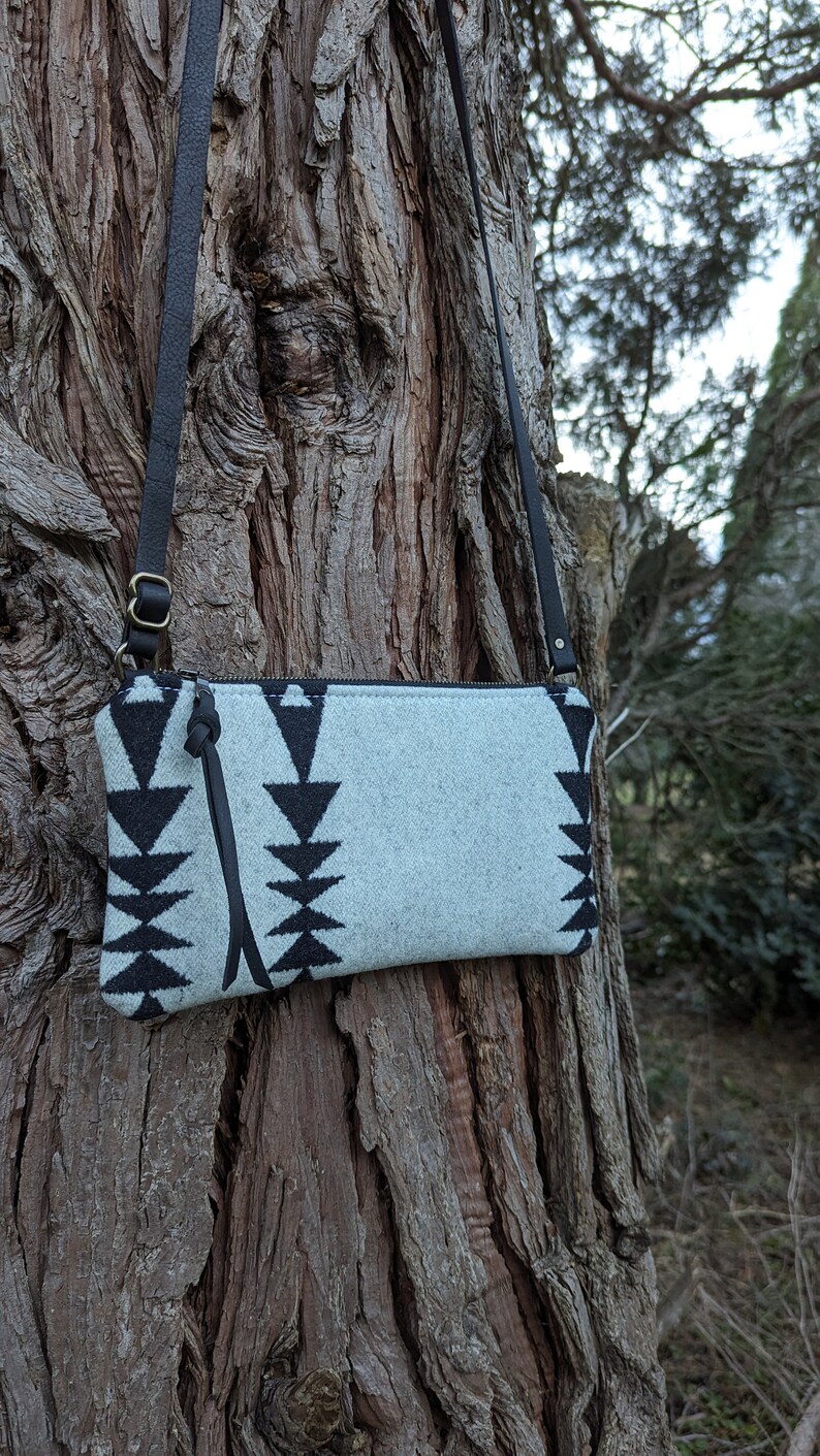 Puede incluir: Un bolso bandolera azul claro con un patr&oacute;n geom&eacute;trico negro. El bolso tiene una correa de cuero negro y un tirador de cremallera. El bolso est&aacute; colgado de un tronco de &aacute;rbol. El bolso mide aproximadamente 25 cm de ancho y 15 cm de alto.