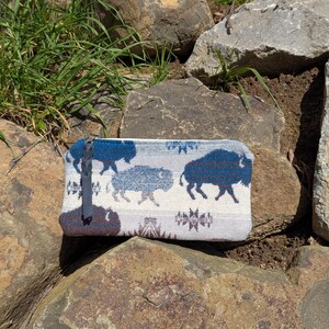 Puede incluir: Bolsa rectangular con un patr&oacute;n repetido de siluetas de bisonte azul sobre un fondo gris claro. La bolsa, de unos 20 cm de largo, tiene un tirador de cremallera azul oscuro y descansa sobre rocas texturizadas.