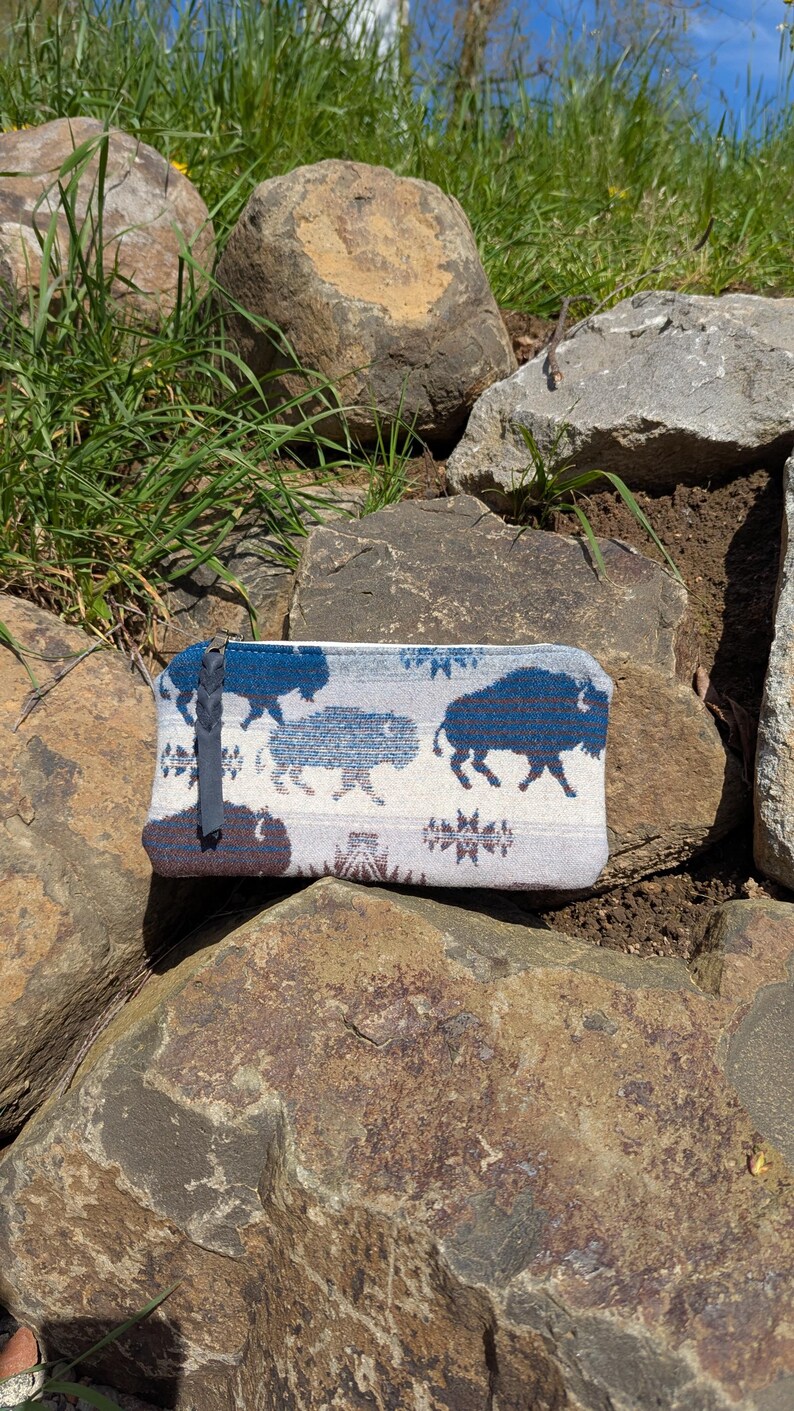 Puede incluir: Bolsa rectangular con un patr&oacute;n repetido de siluetas de bisonte azul sobre un fondo gris claro. La bolsa, de unos 20 cm de largo, tiene un tirador de cremallera azul oscuro y descansa sobre rocas texturizadas.