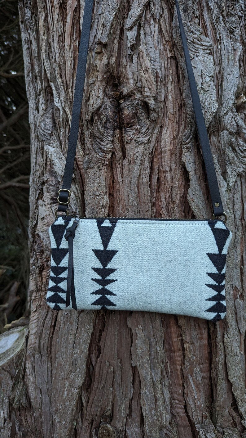 Puede incluir: Un bolso bandolera rectangular con un patr&oacute;n geom&eacute;trico gris claro y negro. El bolso tiene una correa de cuero negro y un tirador de cremallera. El bolso cuelga contra un tronco de &aacute;rbol texturizado.