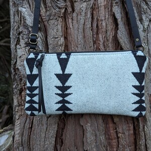 Puede incluir: Un bolso bandolera gris claro con un patr&oacute;n geom&eacute;trico negro. El bolso tiene una correa de cuero negro y cierre de cremallera. El bolso mide aproximadamente 25 cm de ancho y 15 cm de alto.