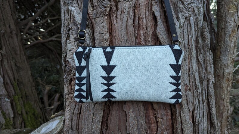 Puede incluir: Un bolso bandolera gris claro con un patr&oacute;n geom&eacute;trico negro. El bolso tiene una correa de cuero negro y cierre de cremallera. El bolso mide aproximadamente 25 cm de ancho y 15 cm de alto.