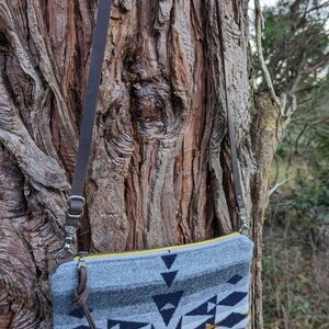 May include: A gray and navy blue patterned crossbody bag with a yellow zipper and brown leather strap. The bag features a geometric design with black and gold accents. The bag is hanging on a tree trunk.