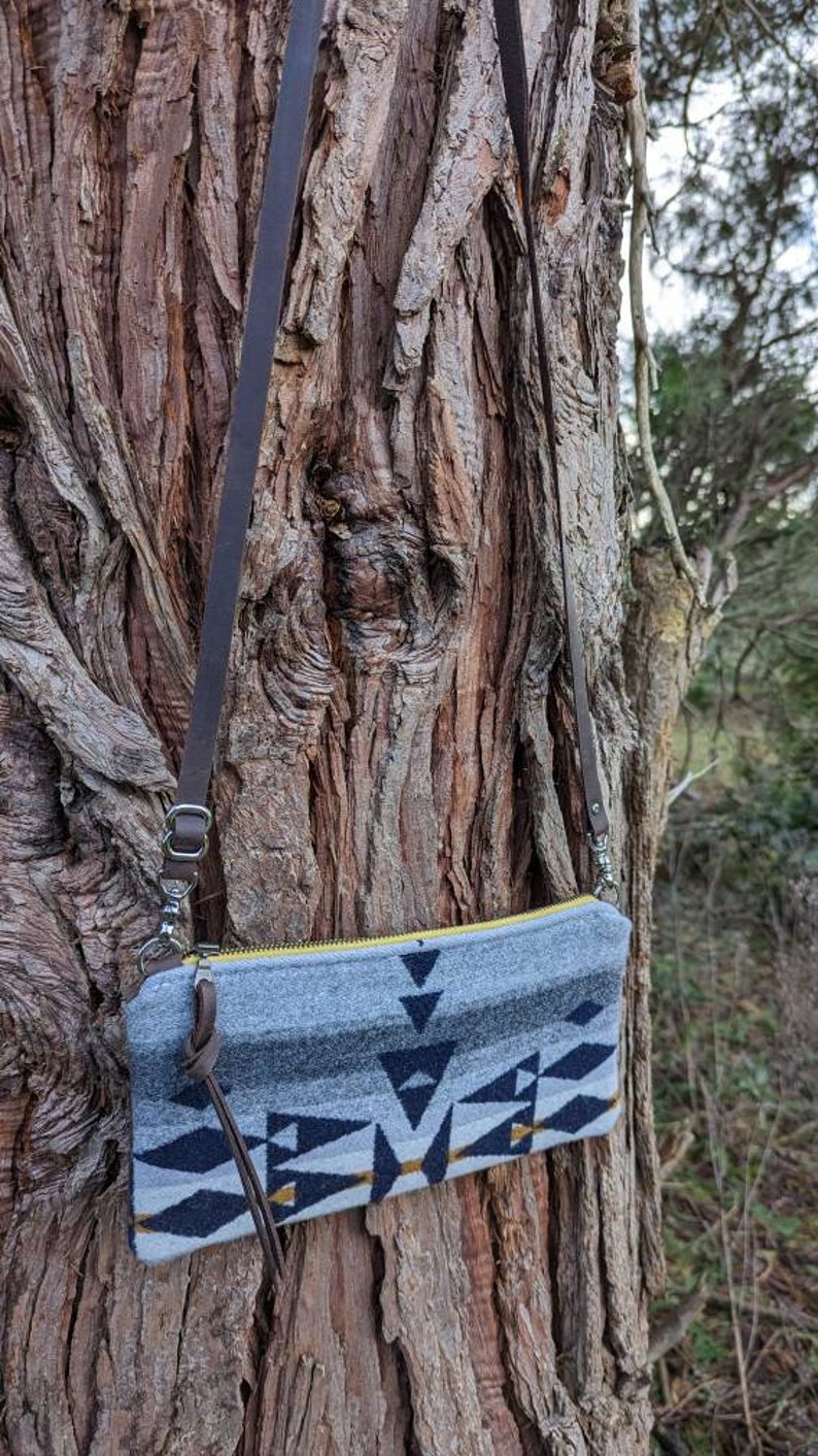 May include: A gray and navy blue patterned crossbody bag with a yellow zipper and brown leather strap. The bag features a geometric design with black and gold accents. The bag is hanging on a tree trunk.