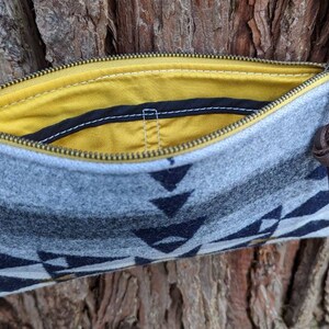 May include: A gray and navy blue patterned crossbody bag with a yellow interior. The bag has a zipper closure, brown leather accents, and a brown leather strap. The bag is made of a soft, felt-like material.