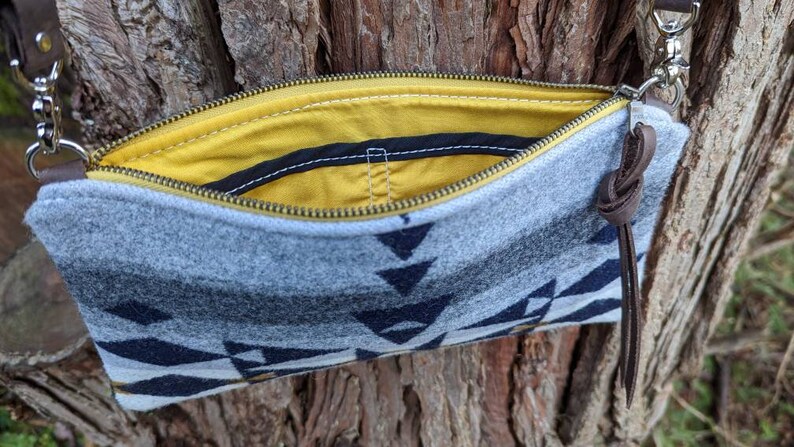 May include: A gray and navy blue patterned crossbody bag with a yellow interior. The bag has a zipper closure, brown leather accents, and a brown leather strap. The bag is made of a soft, felt-like material.