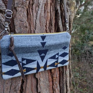 May include: A gray and navy blue crossbody bag with a geometric pattern and a yellow zipper. The bag has a brown leather strap and is hanging on a tree trunk. The bag's design includes triangles and diamond shapes.