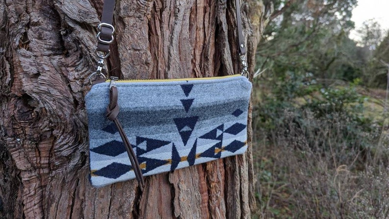 May include: A gray and navy blue crossbody bag with a geometric pattern and a yellow zipper. The bag has a brown leather strap and is hanging on a tree trunk. The bag's design includes triangles and diamond shapes.