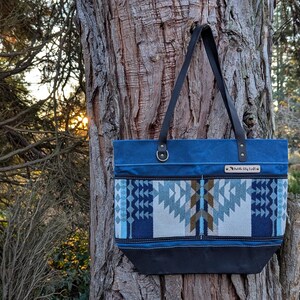 May include: A blue tote bag with a geometric pattern in shades of blue, white, and brown. The bag has black leather handles and a black base. It is hanging on a tree trunk.