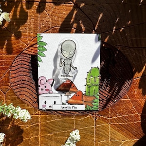 Peut inclure: Une épingle en acrylique transparente représentant un extraterrestre de dessin animé faisant un signe de paix. L'épingle est entourée d'autres personnages de dessin animé, dont un champignon, un cactus et un cochon rose. Le texte "Weird Vibes Only" et "Acrylic Pin" est imprimé sur la carte.