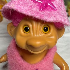 Könnte beinhalten: Eine rosafarbene Trollpuppe mit einem rosafarbenen Hut mit einem lila Seestern darauf. Die Puppe trägt ein rosafarbenes Kleid mit einem lila Seestern darauf.