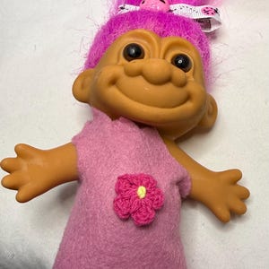 Könnte beinhalten: Eine Troll-Puppe mit leuchtend pinkem Haar und gebräunter Haut, bekleidet mit einem pinken Filzkleid mit gezacktem Saum. Eine kleine pinke Häkelblume mit gelber Mitte ist am Kleid befestigt. Eine weiße Haarspange mit Marienkäfer-Muster ist sichtbar.