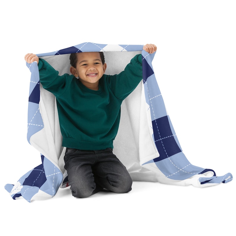 May include: A light blue and navy argyle patterned blanket. The blanket is held up by a child wearing a green sweater and dark gray pants. The blanket has a white backing.