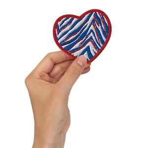 May include: Embroidered heart-shaped patch with a red border and a blue and white zebra stripe design. The patch is held in a hand against a white background. This patch could be used to decorate clothing or accessories.