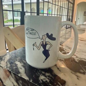 May include: White ceramic mug with a black and white illustration of a woman in a dress and hat. The mug has the text "But I am Le Tired!" in a speech bubble.