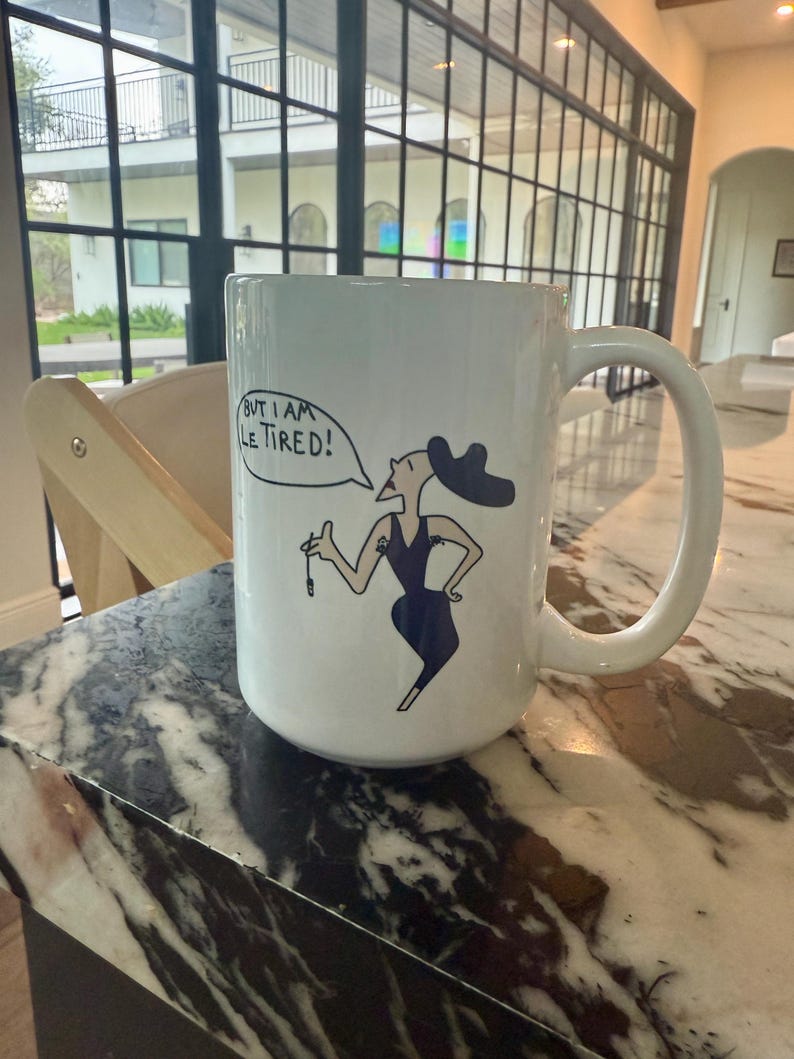 May include: White ceramic mug with a black and white illustration of a woman in a dress and hat. The mug has the text "But I am Le Tired!" in a speech bubble.
