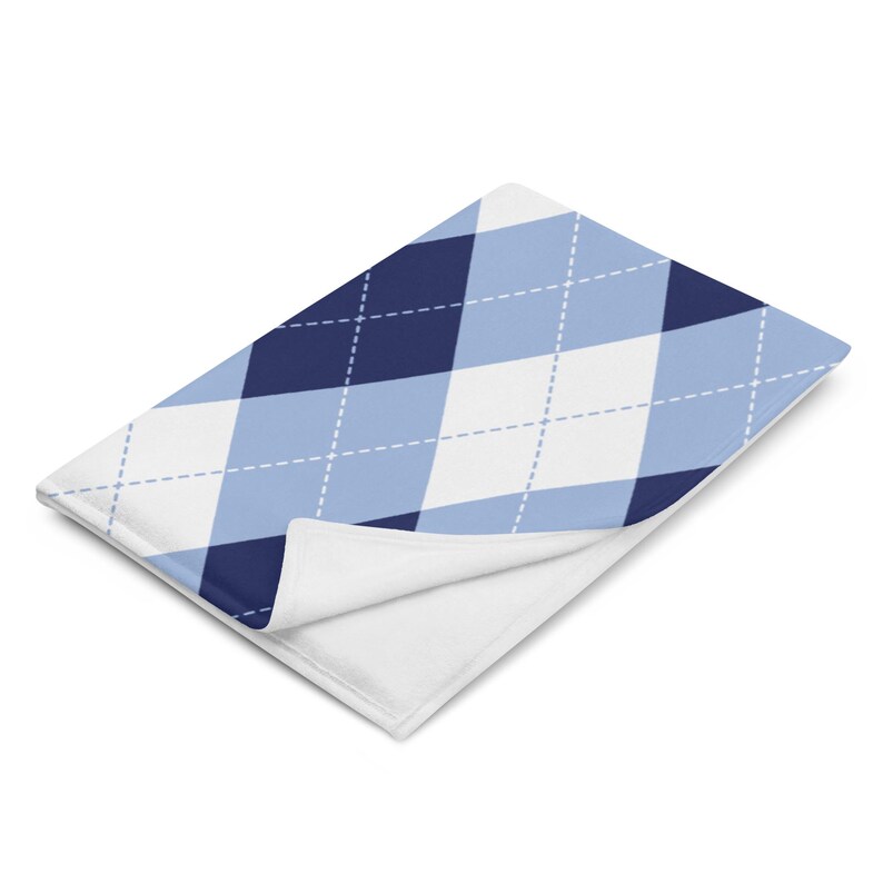 May include: A folded blanket featuring a classic argyle pattern in shades of navy blue, light blue, and white. The blanket has a soft, plush texture and a white underside. The argyle design is composed of diamond shapes with dashed lines.