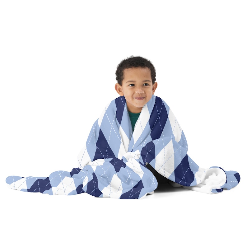 May include: A baby blanket with a blue, white, and navy argyle pattern. The blanket is wrapped around a child. The blanket has a soft, quilted texture and is designed for comfort and warmth.