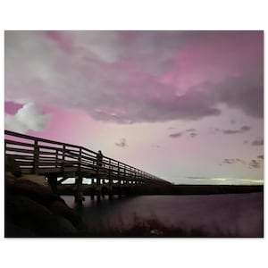 Może przedstawiać: Drewniany pomost rozciąga się nad wodą, a na jego końcu stoi osoba patrząca na różowe i fioletowe niebo. Niebo jest pełne chmur, tworząc dramatyczną i kolorową scenę.