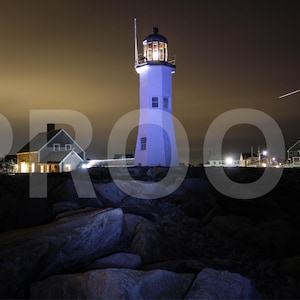 May include: A white lighthouse with a black roof and a light on top, illuminated at night. The lighthouse is on a rocky shore with a small building in the background.