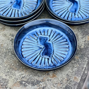 May include: Set of four blue ceramic coasters featuring a raised outline of the state of New Jersey. Each coaster has a sunburst design radiating from the state outline.