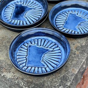 May include: Set of six blue ceramic coasters featuring the outline of the state of Minnesota. Each coaster has a sunburst design radiating from the center.