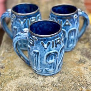 May include: Three blue ceramic mugs with handles. The mugs have a textured surface and feature the words "S VIRGINIA 1972" in raised lettering. The interior of the mugs is a darker blue, and the rims are a darker color.