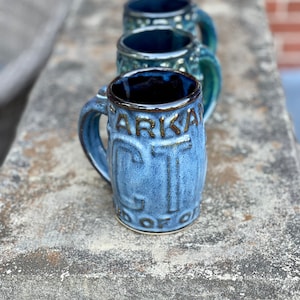 May include: Three blue ceramic mugs with a dark blue glaze and brown accents. The mug in the foreground has the words "ARIKA CITY" embossed on the side.