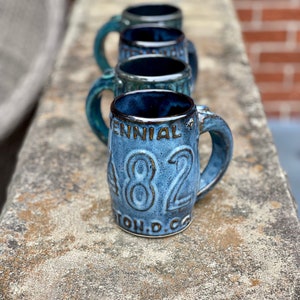 May include: A row of five blue ceramic mugs with a white glaze. The mug in the foreground has the text "Centennial 1882" and "Town, D.C." on it.