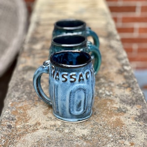 May include: Three blue ceramic mugs with a textured surface. The mug in the foreground has the word "MASSACHUSETTS" written on it.