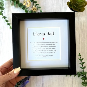 May include: A framed print with the text "Like-a-dad" and a touching message. The black frame surrounds a white mount, with the text in a simple font. A small red heart is above the message. The frame is held by a person.