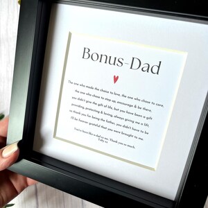 May include: A framed print with the words "Bonus-Dad" at the top, followed by a heartfelt message. The black frame surrounds a white mat and the printed text. A small red heart is centered above the message.