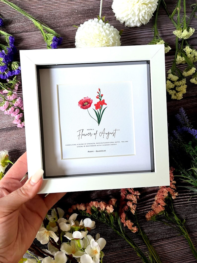May include: A white framed print with a red poppy and a pink gladiolus flower. The text reads "You're a Flower of August. Symbolizing a blend of strength, passion & emotional depth. You are strong & independent with a vibrant energy. Poppy - Gladiolus"