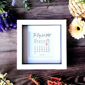 May include: A white framed print with the text "The Day We Met" and a calendar for July 2023 with a pink heart marking the 7th. The text "Cory & Ryan" is at the bottom of the calendar.