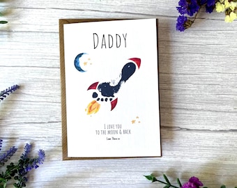 May include: A greeting card with the word "DADDY" at the top. Below is a rocket ship design made with a footprint, with the text "I LOVE YOU TO THE MOON & BACK" and a name. The card is surrounded by flowers.