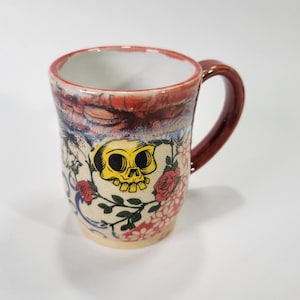 May include: A ceramic mug with a red handle and a white background. The mug is decorated with a yellow skull, red roses, and other floral designs.