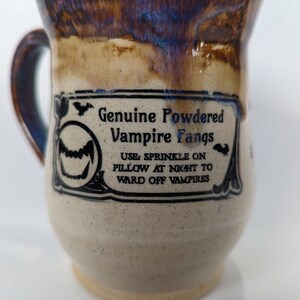 May include: A brown and white ceramic mug with a black and white label that reads "Genuine Powdered Vampire Fangs. Use: Sprinkle on pillow at night to ward off vampires."