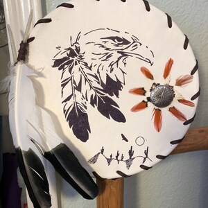 May include: Round, off-white leather shield with a hand-drawn eagle and feather design. The shield features a brown stitched border, a silver button, and attached feathers. The design includes a desert landscape with cacti and a flying bird.