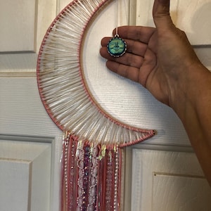 May include: A pink crescent moon dream catcher with white yarn and pink, white, and iridescent beads hanging from the bottom. A small, round, green and blue glass charm with a face is hanging from the top of the dream catcher.