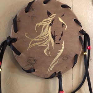 May include: A round leather dreamcatcher with a gold horse silhouette design. The dreamcatcher has black leather ties with red beads.