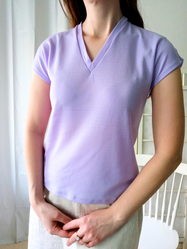 T-shirt vintage neuf en deadstock Ban-Lon années 60. Le T-shirt est de couleur violette, avec un col en V élégant, évoquant la mode rétro de cette époque. La coupe est adaptée pour les tailles S/M (36/38) ou M/L (38/40), mettant en valeur un look décontracté et sportif. Son style reflète l’esprit libre et la mode iconique des années 60, avec une touche moderne grâce à la fraîcheur du vêtement neuf et authentique.