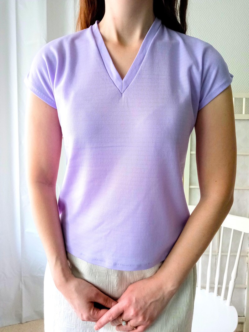 T-shirt vintage neuf en deadstock Ban-Lon années 60. Le T-shirt est de couleur violette, avec un col en V élégant, évoquant la mode rétro de cette époque. La coupe est adaptée pour les tailles S/M (36/38) ou M/L (38/40), mettant en valeur un look décontracté et sportif. Son style reflète l’esprit libre et la mode iconique des années 60, avec une touche moderne grâce à la fraîcheur du vêtement neuf et authentique.