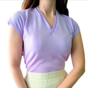 T-shirt vintage neuf en deadstock Ban-Lon années 60. Le T-shirt est de couleur violette, avec un col en V élégant, évoquant la mode rétro de cette époque. La coupe est adaptée pour les tailles S/M (36/38) ou M/L (38/40), mettant en valeur un look décontracté et sportif. Son style reflète l’esprit libre et la mode iconique des années 60, avec une touche moderne grâce à la fraîcheur du vêtement neuf et authentique.