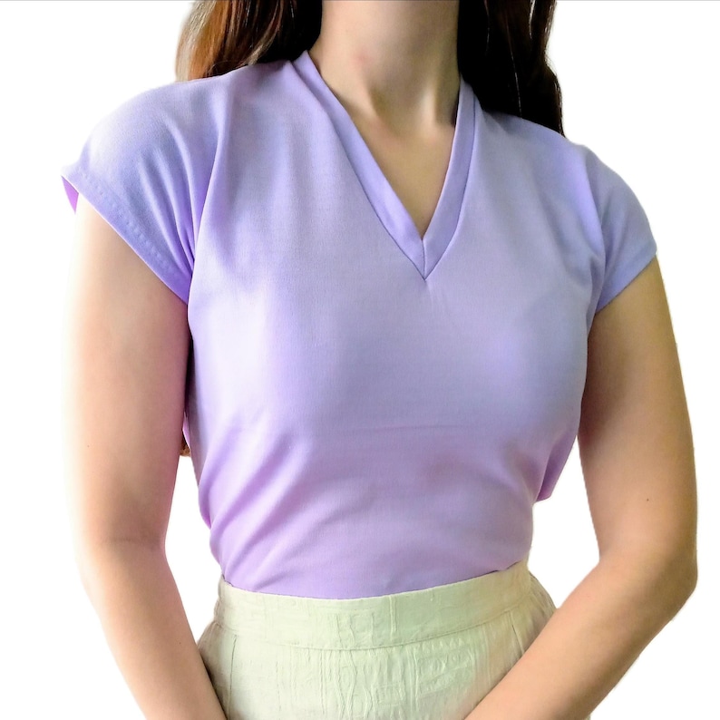T-shirt vintage neuf en deadstock Ban-Lon années 60. Le T-shirt est de couleur violette, avec un col en V élégant, évoquant la mode rétro de cette époque. La coupe est adaptée pour les tailles S/M (36/38) ou M/L (38/40), mettant en valeur un look décontracté et sportif. Son style reflète l’esprit libre et la mode iconique des années 60, avec une touche moderne grâce à la fraîcheur du vêtement neuf et authentique.