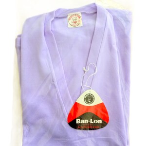 T-shirt vintage neuf en deadstock Ban-Lon années 60. Le T-shirt est de couleur violette, avec un col en V élégant, évoquant la mode rétro de cette époque. La coupe est adaptée pour les tailles S/M (36/38) ou M/L (38/40), mettant en valeur un look décontracté et sportif. Son style reflète l’esprit libre et la mode iconique des années 60, avec une touche moderne grâce à la fraîcheur du vêtement neuf et authentique.