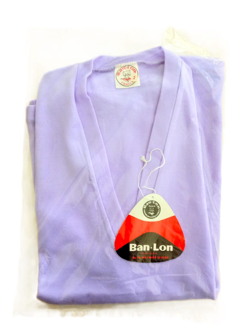 T-shirt vintage neuf en deadstock Ban-Lon années 60. Le T-shirt est de couleur violette, avec un col en V élégant, évoquant la mode rétro de cette époque. La coupe est adaptée pour les tailles S/M (36/38) ou M/L (38/40), mettant en valeur un look décontracté et sportif. Son style reflète l’esprit libre et la mode iconique des années 60, avec une touche moderne grâce à la fraîcheur du vêtement neuf et authentique.