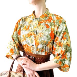 May include: A patterned button-down shirt in orange, green, and yellow. The shirt has short sleeves and is worn with a brown belt and pleated skirt. A woven handbag is held in the hand.