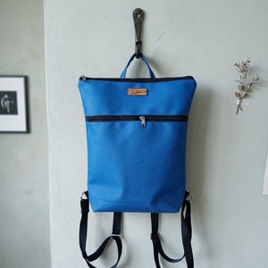 Può includere: Uno zaino blu brillante con cerniere e cinghie nere è appeso a un gancio di metallo. La borsa presenta una piccola etichetta di legno e una maniglia superiore. Il design è semplice e moderno.