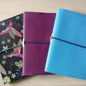 Può includere: Tre diari rilegati in pelle con copertine diverse. Un diario ha una copertina nera con un motivo di colibrì colorato, un altro ha una copertina viola e il terzo ha una copertina blu.